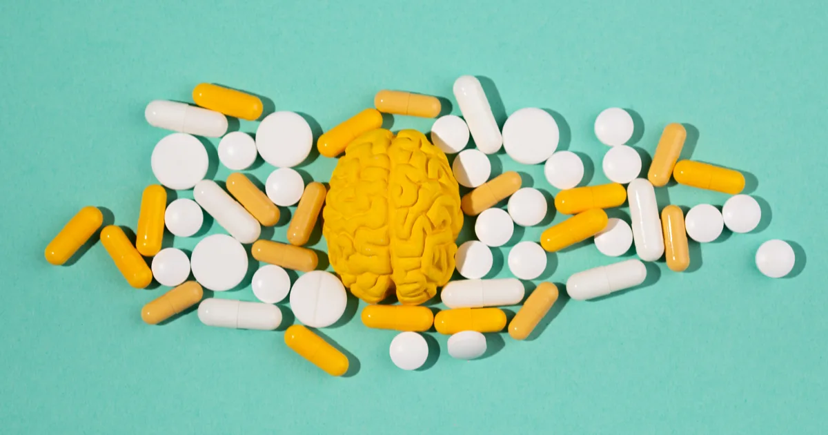A model of a brain surrounded by yellow and white pills on a turquoise background, representing the treatment and study of neurological symptoms.