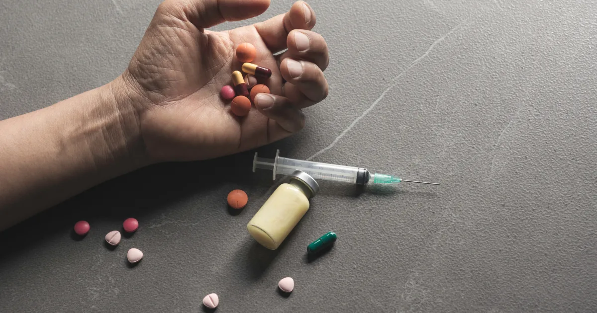 Human hand holding various pills with a syringe and vial nearby, representing medication-related complications such as drug induced movement disorders.