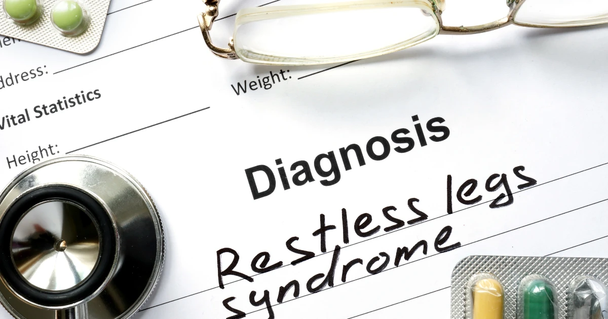 Medical diagnosis form showing “Restless Leg Syndrome” written by hand, with a stethoscope, pills, and eyeglasses placed nearby.