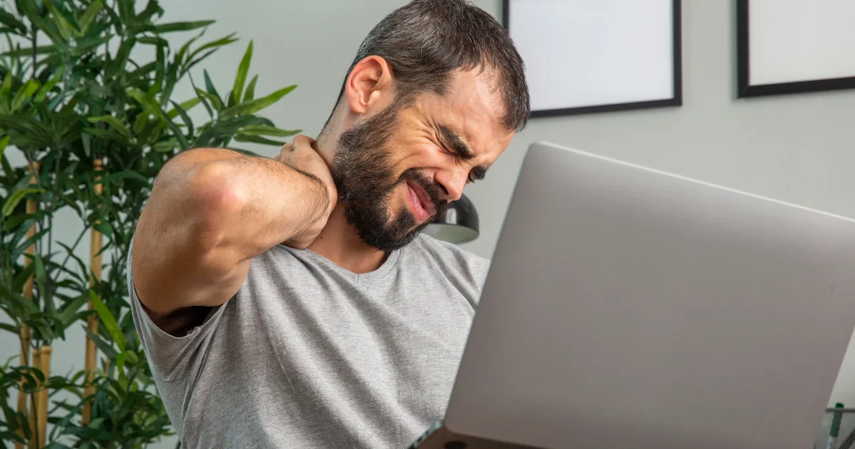 Man experiencing neck stiffness while working on a laptop, representing CBD symptoms such as muscle tightness and jerks.