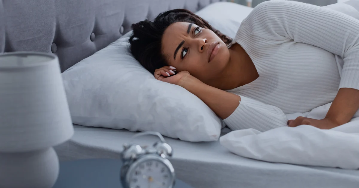 Woman lying awake in bed, looking uncomfortable, possibly after episodes of kicking during sleep linked to REM Sleep Behavior Disorder.