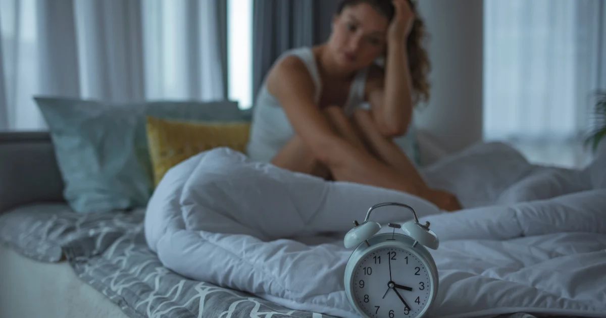 Woman sitting awake in bed at night with an alarm clock in the foreground, showing sleep disturbance related to RBD in women.