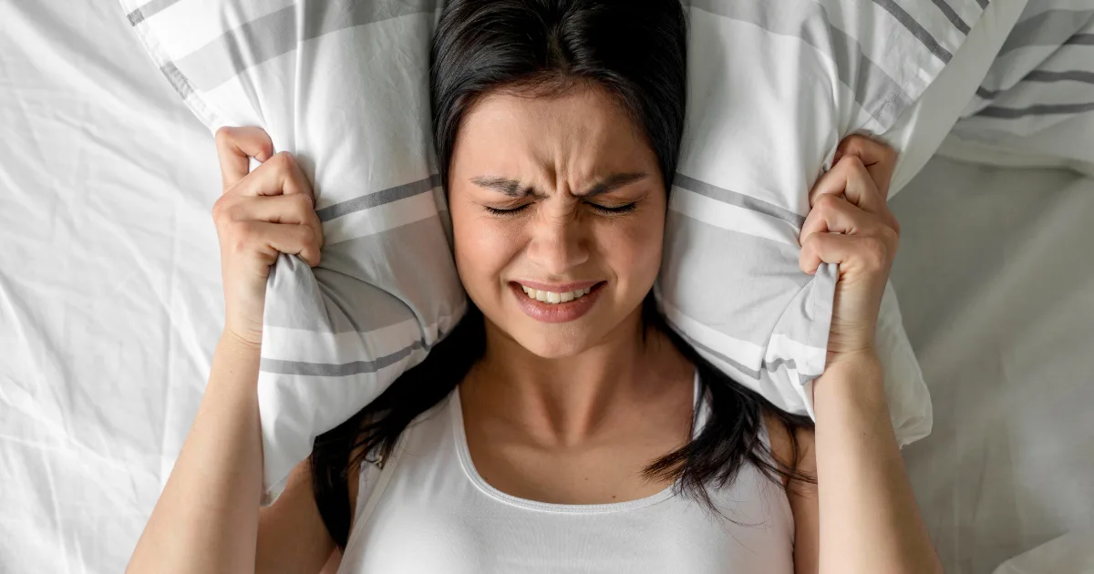 Woman covering her ears with a pillow and looking distressed, reacting to shouting or loud noises during sleep.